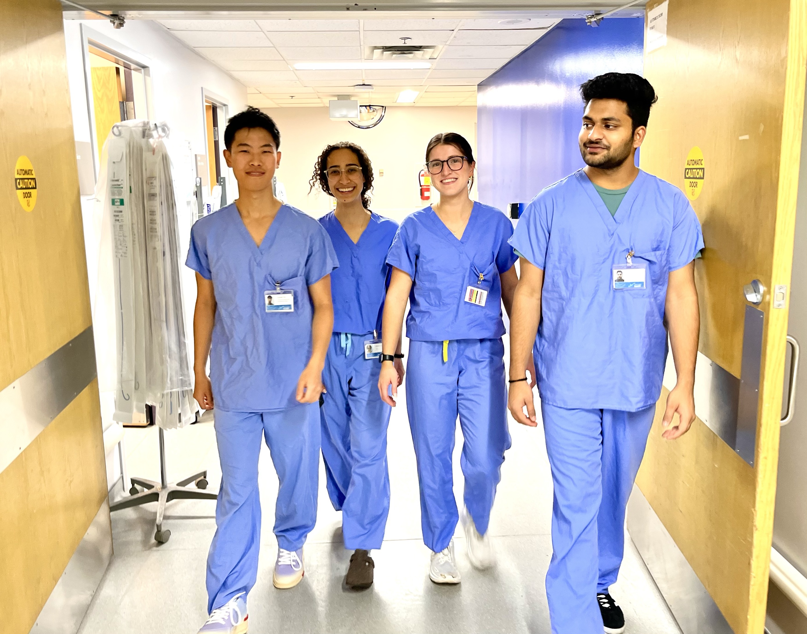 Four students wearing blue scrubs walk through double doors in a hospital hallway. 