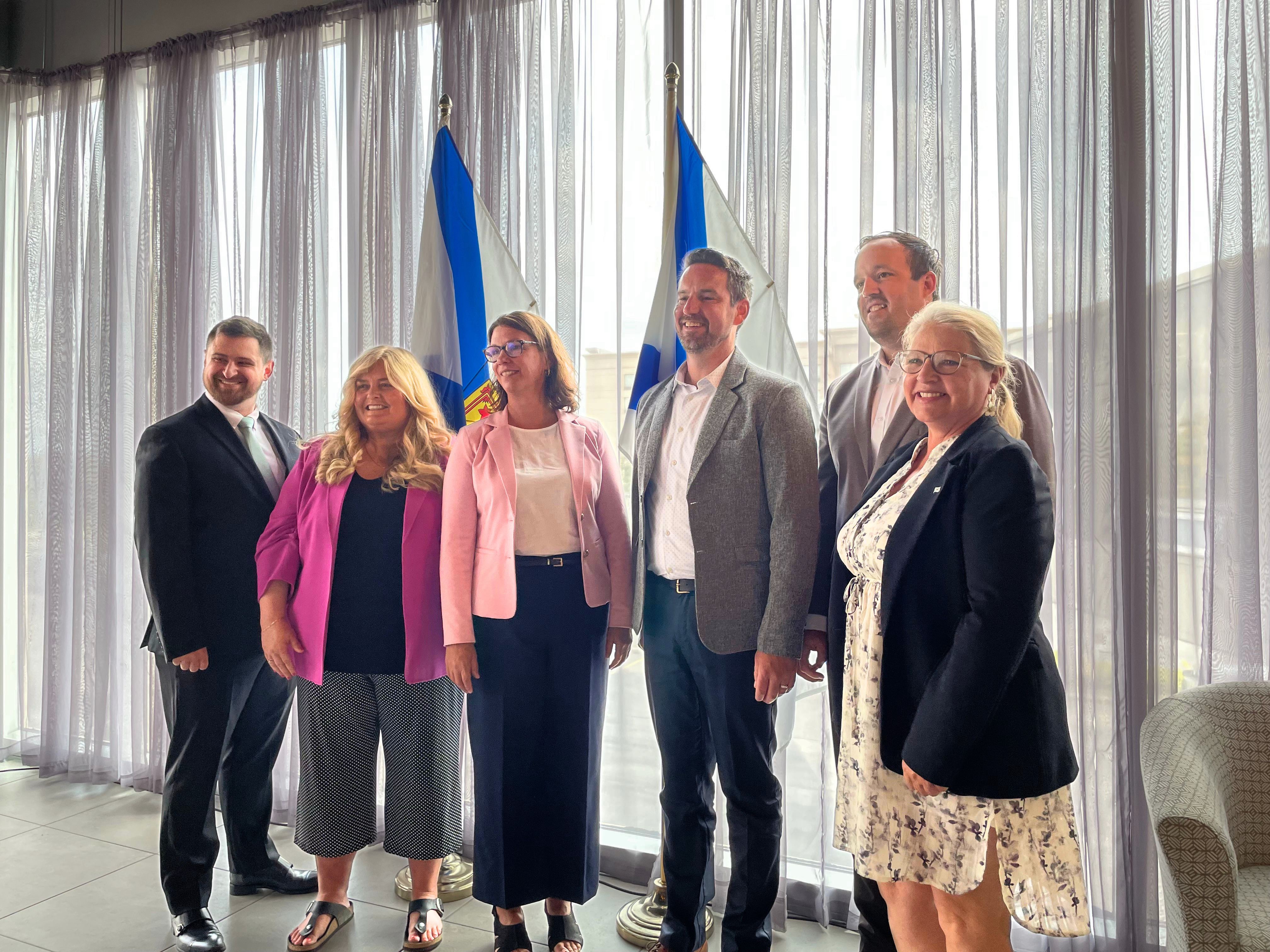 Nova Scotia Health Representatives, Nova Scotia Government Officials, and AstraZeneca Leadership pose in a photo at an announcement celebrating a new cancer centre in Cape Breton. The Nova Scotian flags are pictured behind the group. 