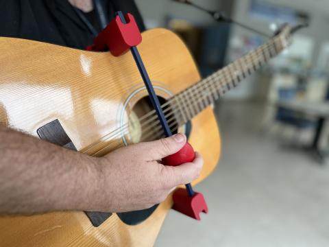 A close up picture of a person's arm and hand holding a guitar that is fitted with StrumAssist, a 3D-printed device to help patients with limited motor function strum. 