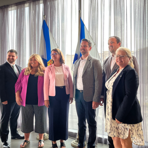 Nova Scotia Health Representatives, Nova Scotia Government Officials, and AstraZeneca Leadership pose in a photo at an announcement celebrating a new cancer centre in Cape Breton. The Nova Scotian flags are pictured behind the group. 